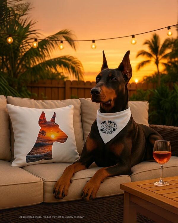 A stylish outdoor patio setup at golden hour—cozy seating with the Sunset Doberman Pillow front and center. A Doberman lounges nearby with a relaxed but alert pose. Background includes string lights, a low sun setting behind palm trees and backyard fence, a glass of rosé nearby. Mood: chill, proud, beautifully Dobie.